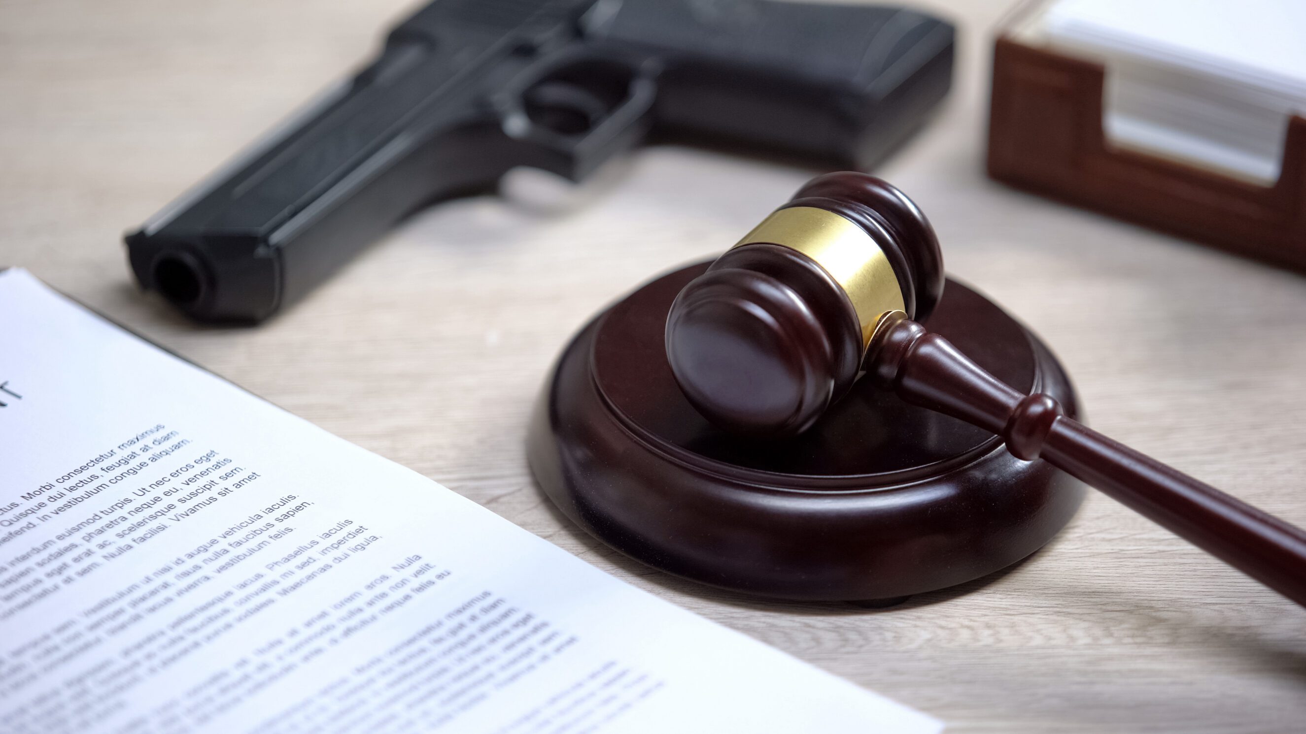 Wooden judge's gavel and sound block on a desk beside estate/legal documents with a handgun partially visible in the background, representing firearm handling and legal oversight for estate and trust matters.