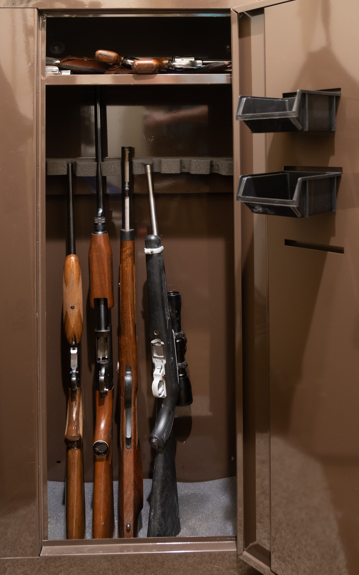 A brown gun safe with its door open showing four long guns with wood and black stocks standing upright, a top shelf holding a leather shotgun case, and two plastic bins mounted on the inside of the door, suggesting secured estate firearms storage and collection handling.